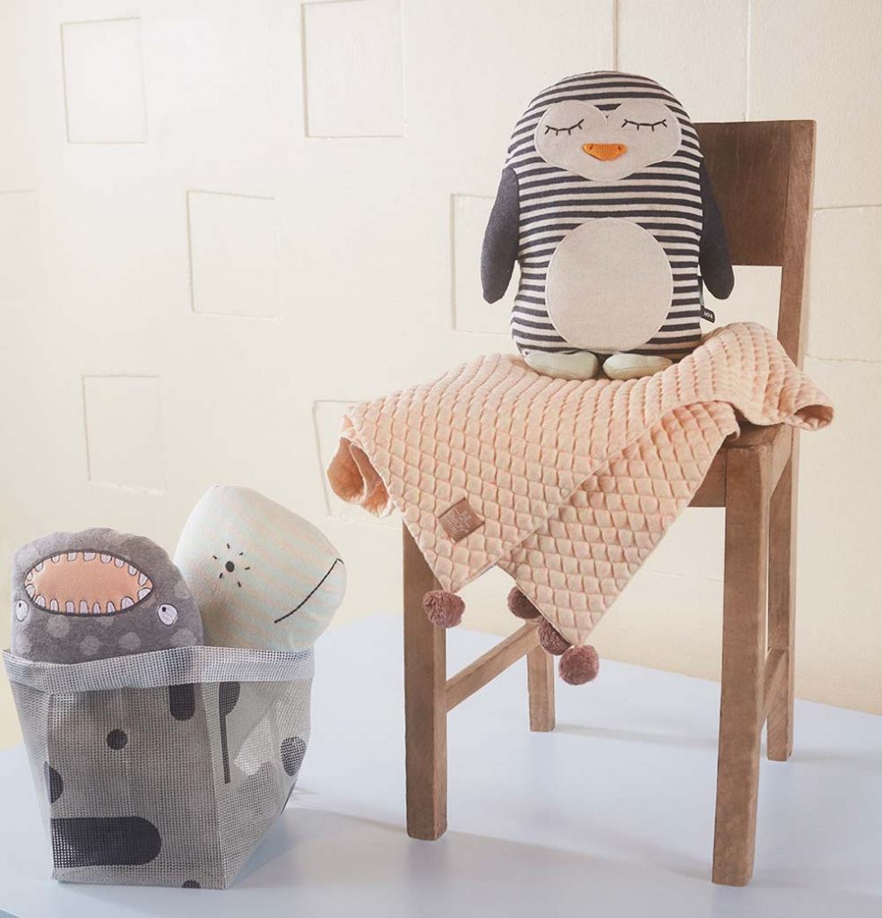 Das Deko-Strickkissen "Pinguin Pingo" dient auch als Kuscheltier und macht dem Baby vor, wie man gut die Augen zumachen kann. Pingo sitzt auf einem Holzstuhl, auf dem auch eine rosa Decke liegt.
