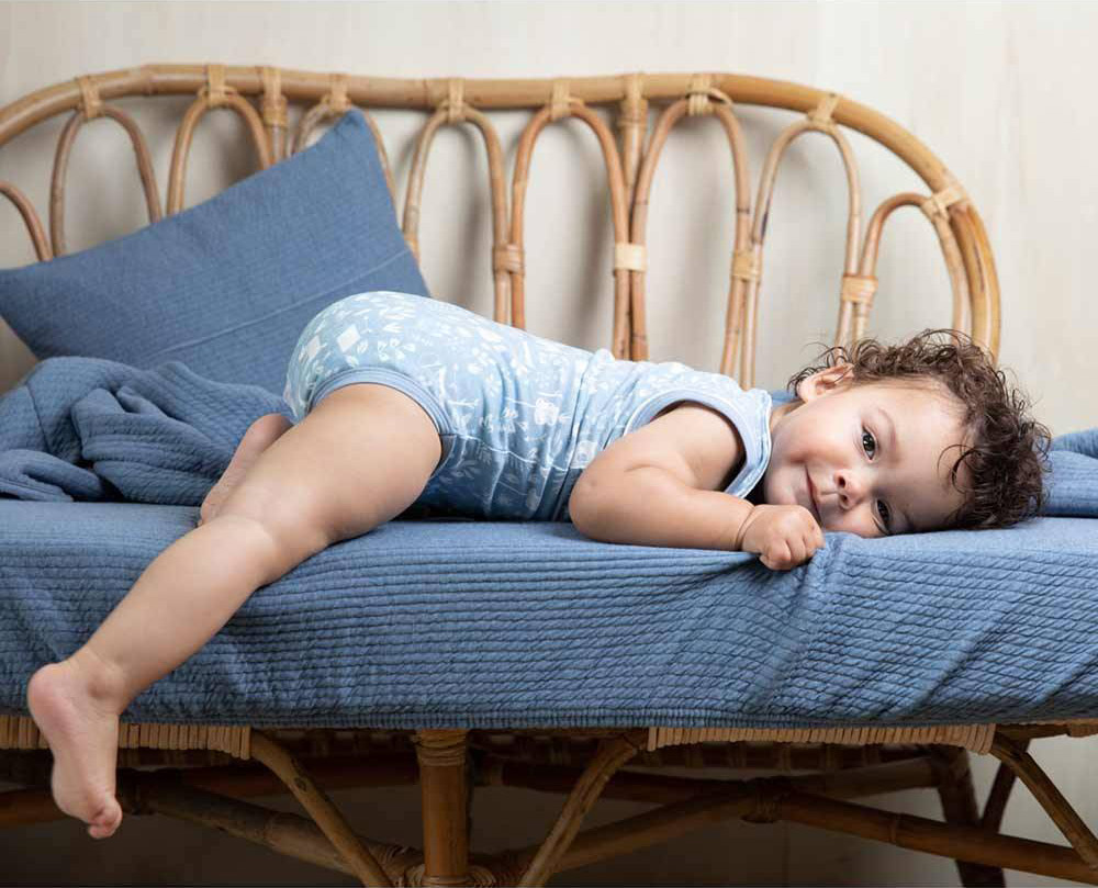 Ein Kleinkind in blauem Body liegt auf einem Rattanbett mit der Little Dutch-Kinderbettwäsche in "Pure Blue" mit den Maßen 100 x 135.