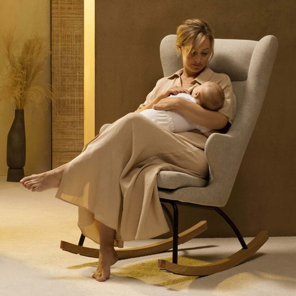 Im Schaukel- und Stillstuhl de Luxe von der Firma Quax in Clay (Beige/Grau) sitzt eine Mutter und wiegt ihr Baby in den Schlaf. Die Umgebung sieht ruhig und natürlich aus.